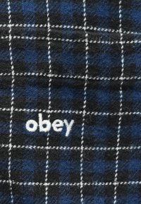 Sinine ja must ruuduline kangas, millel on pehme tekstuur, valge õmblusega ning silmatorkavalt broderitud sõna "obey".
