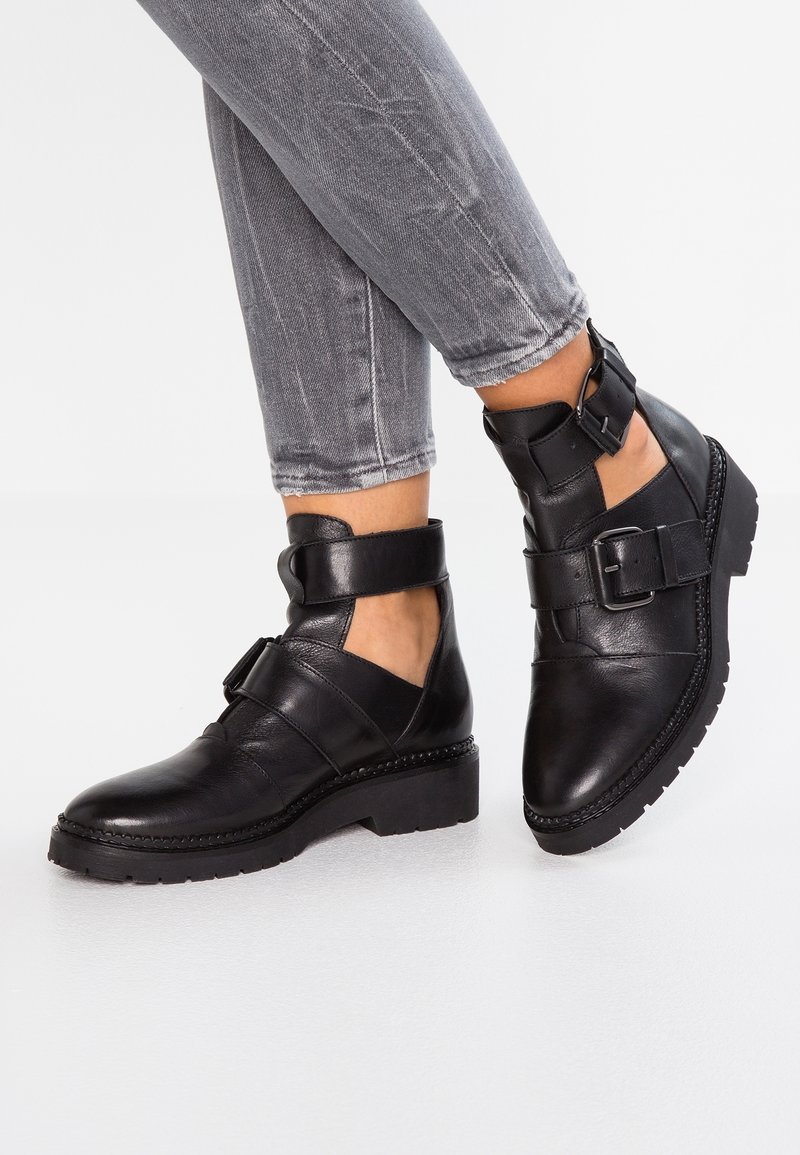 Personne portant des bottines noires à découpes, semelles épaisses et boucles, assorties à un jean gris retroussé, debout sur une surface blanche.