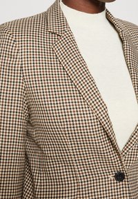 Brązowo-beżowa marynarka w wzór pepitki, z wyciętym kołnierzem i pojedynczym czarnym guzikiem, noszona na jasnym golfie.