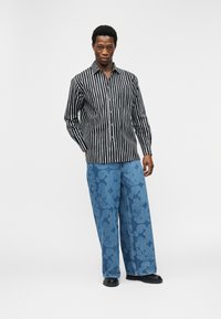 Άνδρας που στέκεται με τα χέρια στις τσέπες, φοράει πουκάμισο με μαύρες και λευκές κάθετες ρίγες και φαρδιά μπλε τζιν με ανθικό σχέδιο.