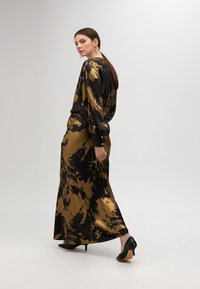 Lange, zwart en goud gekleurde tie-dye jurk met lange mouwen, getailleerd silhouet en knopen bij de manchetten. Gecombineerd met zwarte hoge hakken.