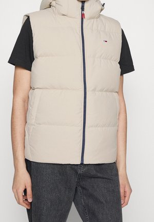 Veste sans manches matelassée beige avec fermeture éclair à l'avant, portée sur un t-shirt noir à manches courtes et un jean gris foncé.