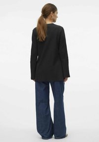 Svart långärmad kappa med rak skärning, kombinerad med blå vida jeans. Slät tygstruktur, minimal detaljer och klassisk design.
