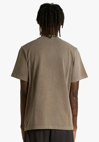 Maglietta marrone in cotone con vestibilità comoda, maniche corte e collo rotondo, caratterizzata da una texture liscia e senza grafiche o motivi.