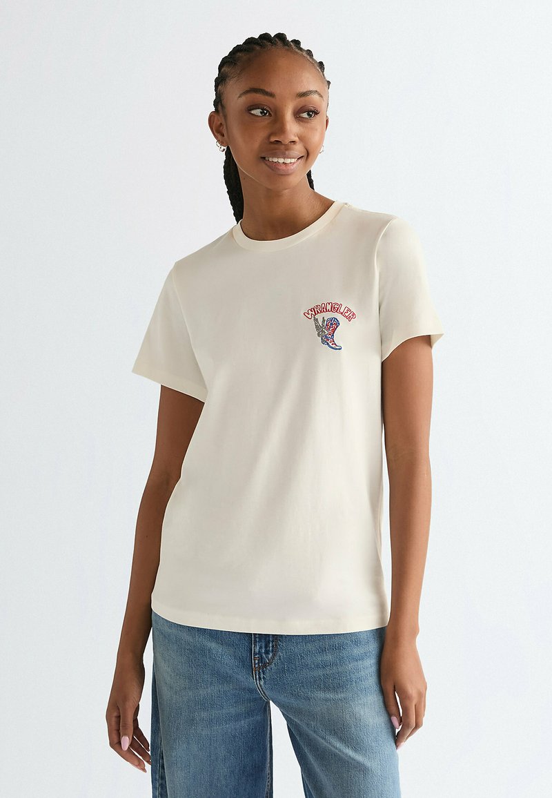 Jonge vrouw met gevlochten haar, draagt een witte Wrangler T-shirt met een klein kleurrijk logo en een blauwe spijkerbroek, glimlacht en kijkt naar rechts.