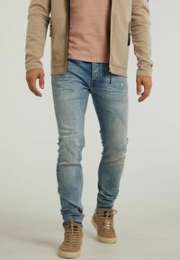Ljust blå denimjeans med bleknade och slitna detaljer, parat med beige mockaskor och en krämfärgad zip-up jacka.