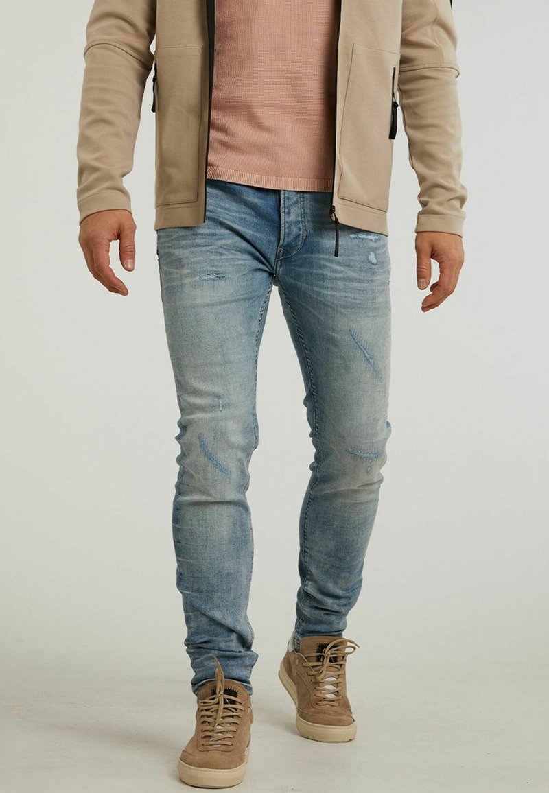 Ljust blå denimjeans med bleknade och slitna detaljer, parat med beige mockaskor och en krämfärgad zip-up jacka.
