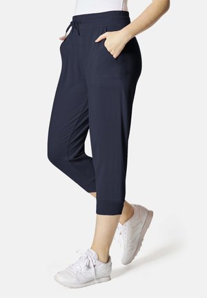 Person trägt navyblaue, verkürzte Hosen mit Taschen und weiße Sneakers, steht mit einer Hand in der Tasche.