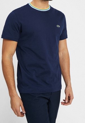 Mężczyzna ubrany w granatowy T-shirt z krótkim rękawem, z wielokolorowym paskowanym okrągłym dekoltem i małym zielonym logo krokodyla na piersi.