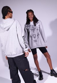 Due persone in felpe grigio chiaro oversize con disegni grafici neri. Una indossa pantaloni neri strappati e stivali neri; l'altra è in pantaloni corti.
