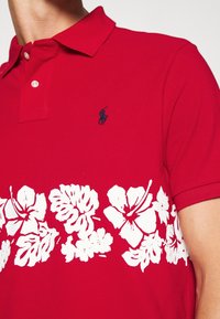 Närbild av en man som bär en röd polo istället med en vit blommönster längst ner och en liten marinblå logo på bröstet.