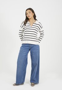Kobieta stoi z rękami za plecami, ubrana w biało-czarny sweter w paski, szerokie niebieskie dżinsy i beżowe baleriny.