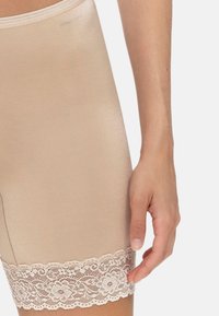Beige formande shorts med blommig spetskant vid låret, bärs av en person med en synlig högerhand som vilar vid benet.