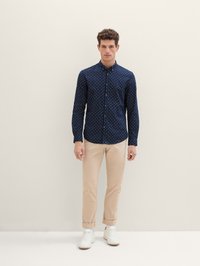 Navy-blaues Hemd mit weißen Punkten, langen Ärmeln und Kragen. Kombiniert mit hellbeigen Chinos und weißen Sneakers. Schlichter Hintergrund.