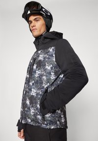 Jachetă de schi cu un corp negru și un front cu model camuflaj în nuanțe de gri și alb; include buzunare cu fermoar și o glugă.