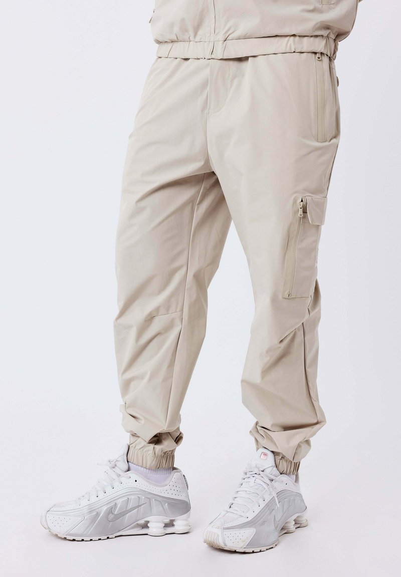 Pantalones cargo beige con puños elásticos, combinados con zapatillas Nike blancas que cuentan con suelas distintivas similares a resortes.