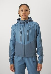 Casaco com capuz e fecho éclair em dois tons de azul, apresentando um design em chevron, punhos elásticos e bolsos no peito com fecho éclair. Superfície suave.