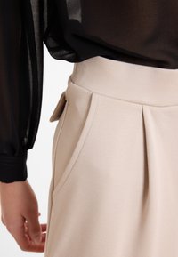 Beige plissé rok van stof met een gladde textuur, gedetailleerde tailleband en zijzakken. Zwarte doorschijnende blouse valt over de top.