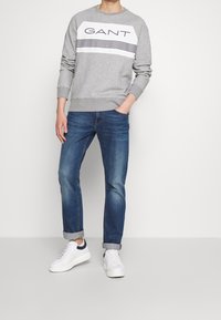 Uomo che indossa un felpa grigia GANT, jeans blu arrotolati e sneakers bianche con dettagli blu navy, in piedi su uno sfondo bianco.