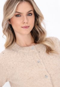 Žena s vlnitými blond vlasy, oblečená v béžovém pleteném svetru zdobeném malými kamínky a dekorativními ozdobnými knoflíky.