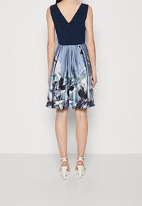 Ärmelloses Kleid mit V-Rücken; einfarbiger navyfarbener Oberkörper und ein ausgestellter Rock mit einem floralen Muster in Blau- und Grüntönen mit Glanz.