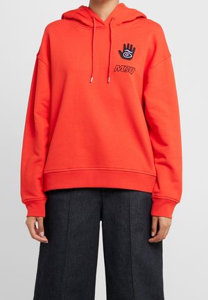 Personne portant un sweat à capuche orange vif avec un symbole noir de main et d'œil et le texte "MCQ" sur la poitrine, associé à un pantalon large foncé.
