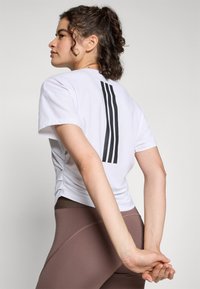T-shirt bianco a maniche corte con strisce verticali nere sul retro, abbinato a leggings marroni. Tessuto morbido con una vestibilità rilassata e una leggera texture.