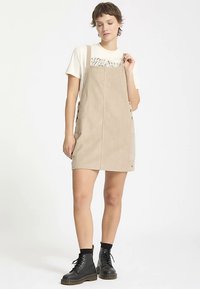Beige corduroy dress with straps, featuring side buttons and a front pocket, paired with a white t-shirt and black lace-up boots.