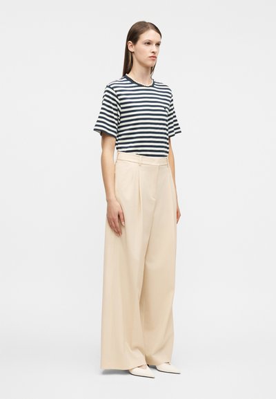 Γυναίκα με μαύρο και άσπρο οριζόντιο ριγέ T-shirt, περιδέραιο με πέρλες, φαρδιά μπεζ παντελόνια και μυτερά λευκά παπούτσια, στέκεται μπροστά σε λευκό φόντο.