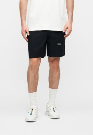 Mann trägt schwarze Shorts mit Fronttaschen, weiße Sportsocken, weiße Turnschuhe und ein weißes T-Shirt, steht vor weißem Hintergrund.