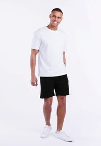 Weißes Baumwoll-T-Shirt mit kurzen Ärmeln, kombiniert mit schwarzen Shorts. Das Model trägt weiße Sneakers und steht vor einem schlichten weißen Hintergrund.