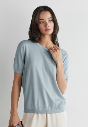 Kvinde med skulderlangt mørkt hår iført en lyseblå kortærmet sweater og cremefarvede bukser, der kigger til siden mod en ensfarvet væg.