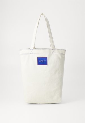 Biała torba typu tote z płótna z dwoma uchwytami oraz niebieską prostokątną naszywką z logo GANT Sportwear na środku z przodu.