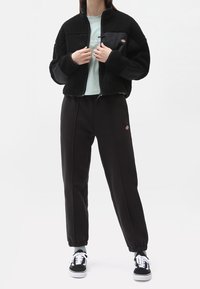 Giacca in pile nera con zip frontale, tessuto testurizzato e tasca laterale; abbinata a pantaloni sportivi neri a gamba affusolata e sneakers nere.