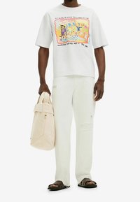Biała koszulka graficzna z kolorową ilustracją, jasne spodnie z detalami przetarcia oraz beżowa torba materiałowa z złożonym zamkiem. Brązowe sandały.
