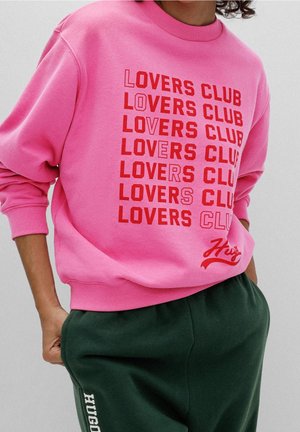 Personne portant un sweat-shirt rose avec le texte rouge répété « LOVERS CLUB » et un pantalon vert foncé avec « HUGO » imprimé verticalement sur la jambe.