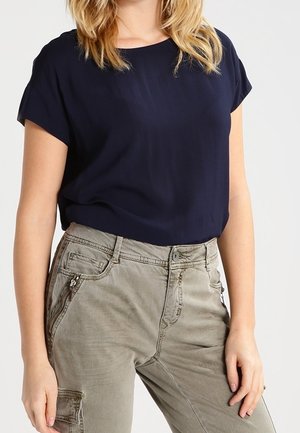Femme portant un haut à manches courtes bleu marine rentré dans un pantalon cargo gris clair avec des poches zippées, debout avec les bras détendus.