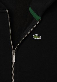 Zwarte hoodie met ritssluiting en een groene voering, voorzien van een metalen rits en een klein geborduurd krokodillenlogo op de borst.