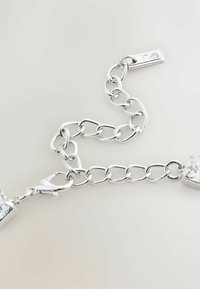 Silberne Kettenarmband mit einem Karabinerverschluss, der an jedem Ende zwei rechteckige klare Kristalle, glatte Metallglieder und ein kleines Etikett aufweist.