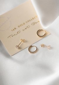 Boucles d'oreilles en or sur carte crème : comprend un clou avec cristal et un design de créole. Texture métallique lisse, finition brillante, accents minimalistes.