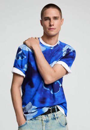 Mann mit kurzen Haaren, der ein blau-weißes Batik-T-Shirt und hellblaue Jeans trägt, posiert mit einer Hand auf der Schulter.