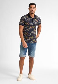 Camisa polo de manga corta con un fondo oscuro que presenta un estampado de palmeras tropicales, combinada con shorts de mezclilla arremangados y zapatillas claras.