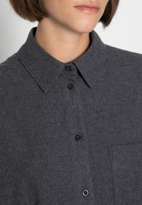 Ciemnoszara koszula z guzikami, wykonana z fakturowanej tkaniny, z kołnierzykiem, czarnymi guzikami i kieszenią na piersi.