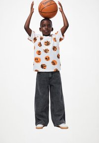 Bela majica s oranžnimi in črnimi vzorci košark in nasmejanih obrazov, v kombinaciji z velikanskimi temno sivimi kavbojkami in belimi supergami.