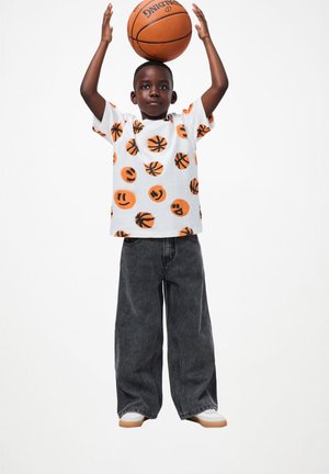 Vit t-shirt med orange och svarta mönster av basketboll och leende ansikten, tillsammans med oversize mörkgrå denimjeans och vita sneakers.