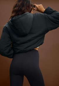 Sort cropped puffer-jakke med hætte, der har en glat tekstur, lagdelt stof og elastiske manchetter, båret med højtaljede sorte leggings.
