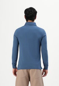 Mann mit dunklen Haaren, der ein blaues Poloshirt mit langen Ärmeln und beigen Hosen trägt, steht mit dem Rücken zu einem schlichten weißen Hintergrund.