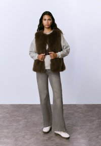 Chaleco de piel sintética marrón sobre una sudadera gris, combinado con jeans de bota acampanada gris claro y zapatillas blancas. Escenario simple con un fondo neutro.