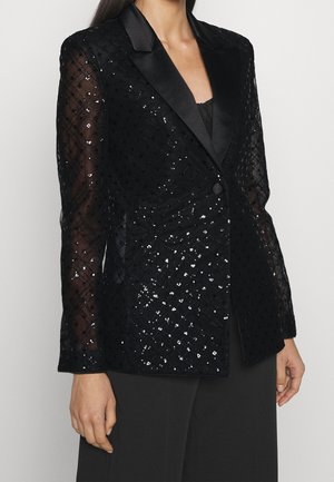 Blazer noir à sequins avec revers satinés et manches transparentes à motifs, porté sur un top et un pantalon noirs.