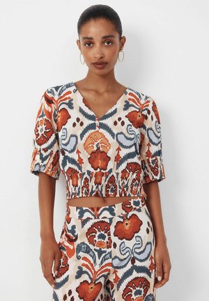 Kvinde iført matchende V-udskåret crop top og højtaljede bukser med orange, marineblåt og beige abstrakt blomstermønster og øreringe i bøjle.